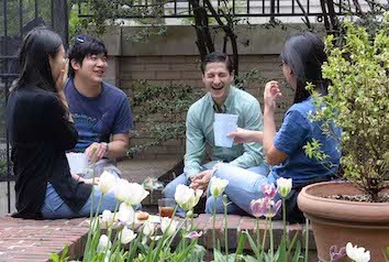 For individuals sitting in a circle, two are facing camera, one is laughing and another is smiling. Flowers in foreground