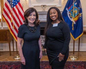 Two women smiling in front of USA flag and New York State flag in background